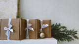 Brown boxed gifts wrapped with white ribbon on a shelf with greenery. Local Gift Guide 2025
