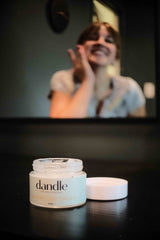 Organic Clarifying face cream in glass jar sitting on a black surface, in the background you can see a girl looking into the mirror applying it