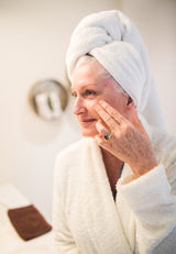 Older woman in a white bathrobe applying skincare to her cheek.