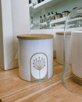 White candle jar with wooden lid on a wooden surface, with shelves of candles in the background.