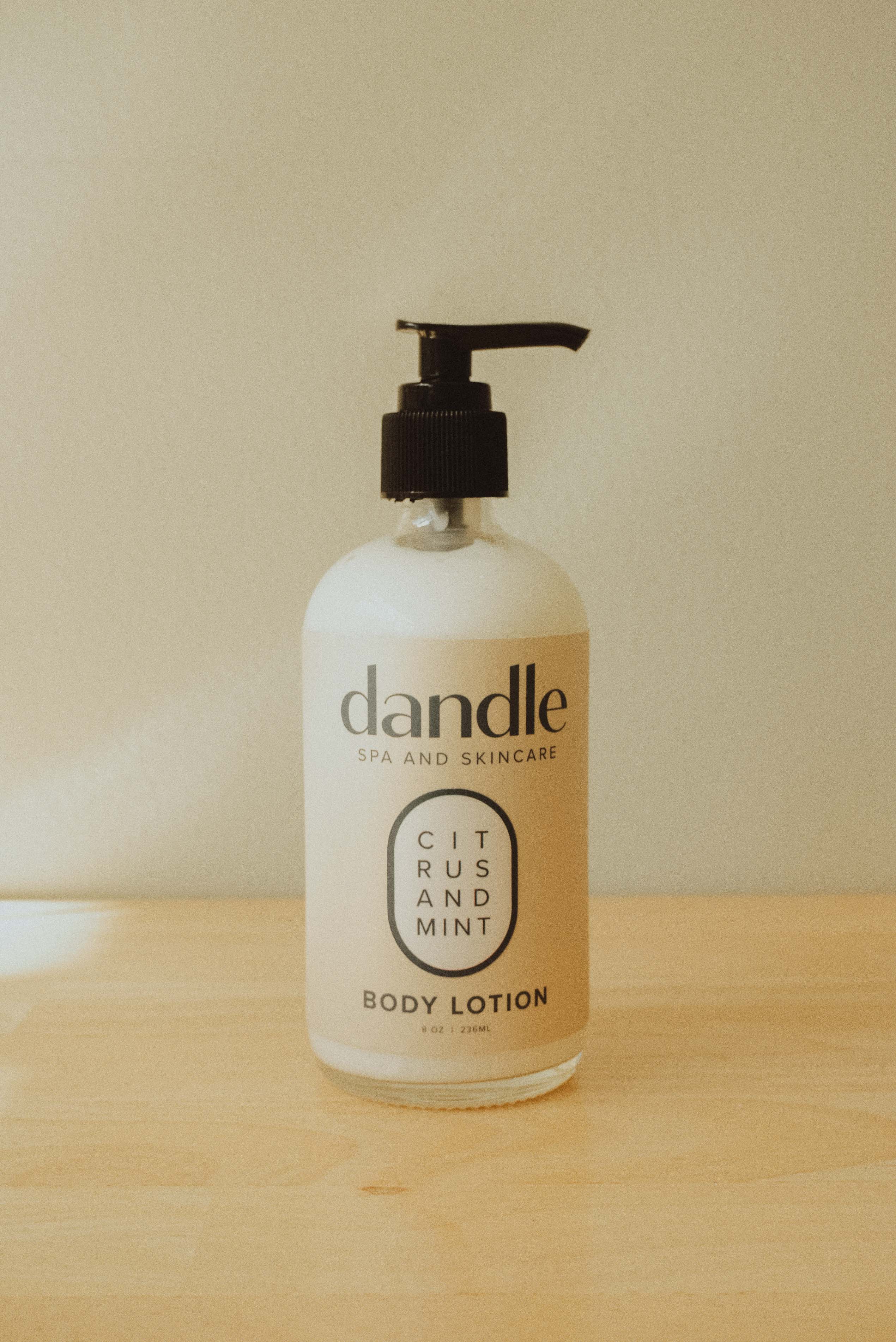 A bottle of Dandle Citrus and Mint Body Lotion on a light wooden surface against a neutral beige background.
