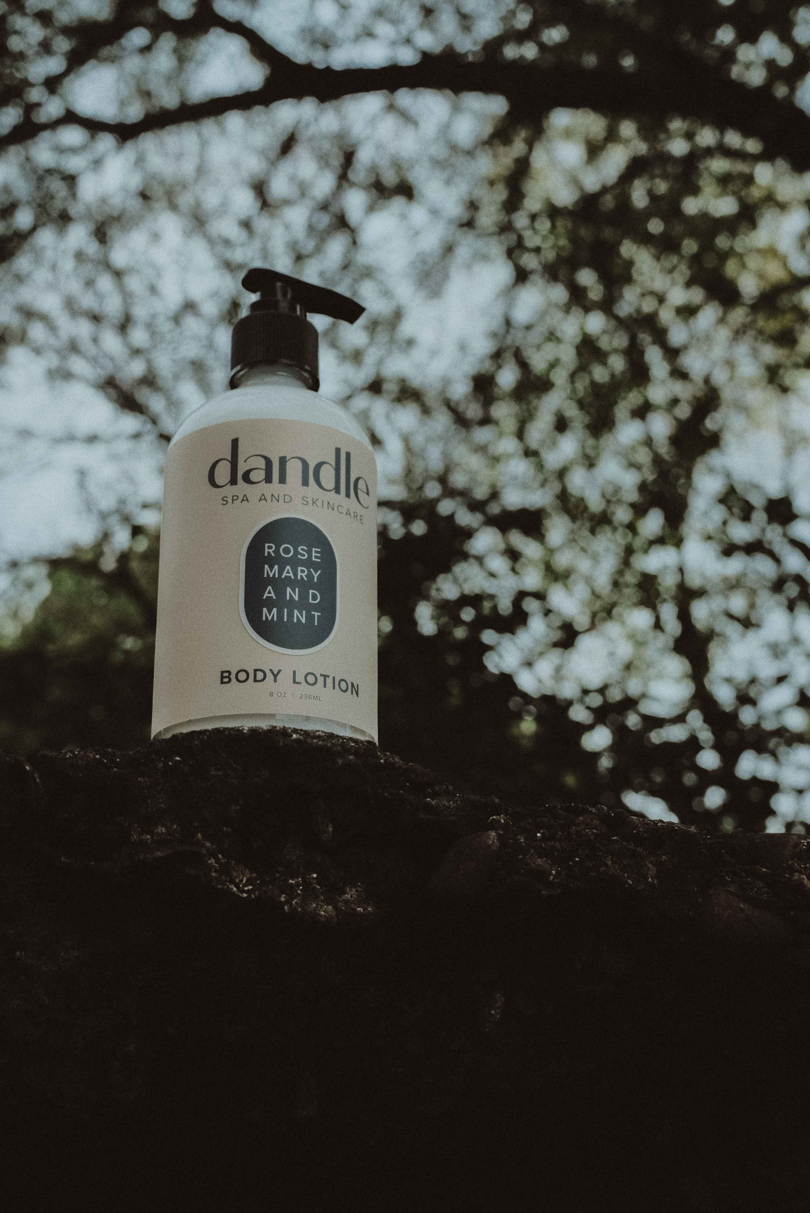 Dandle rosemary mint body lotion bottle on a tree branch with white blossoms in the background. Local Tennessee product. 