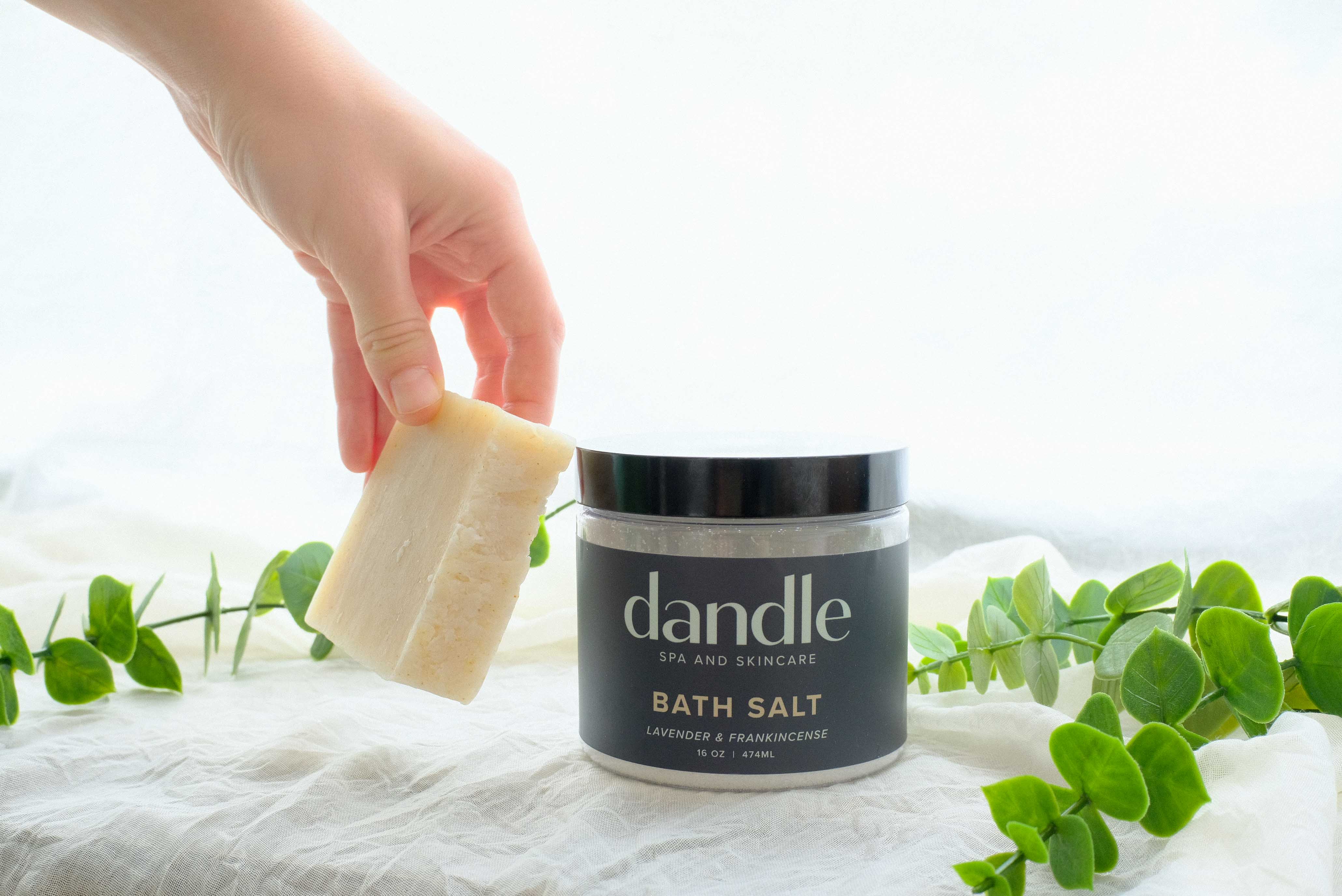 Hand holding a bar of soap next to a jar of dandle bath salts with greenery in the background. Christmas gifts in Chattanooga Tn