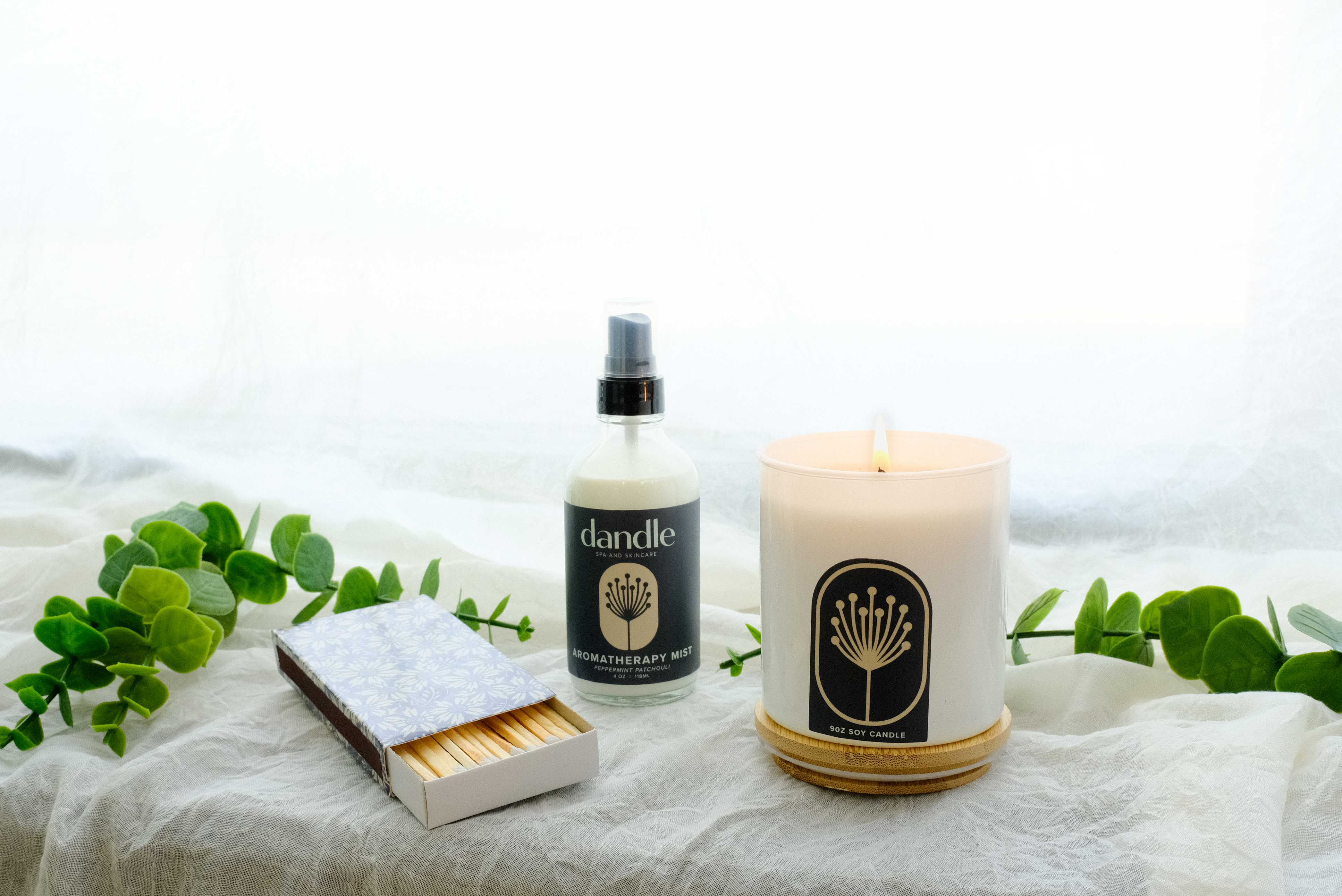 Candle, matches, and  natural room spray on a white surface with green leaves. hand-poured candle made with essential oils. non-toxic candle, all natural room spray, organic linen spray. Locally made Christmas gifts