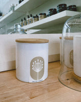 Candle jar with wooden lid on a wooden surface, with shelves in the background containing various items.
