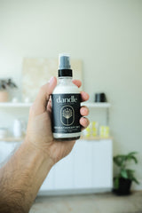 Hand holding a bottle of 'dandle' aromatherapy mist with a blurred indoor background. Organic linen spray for sleep