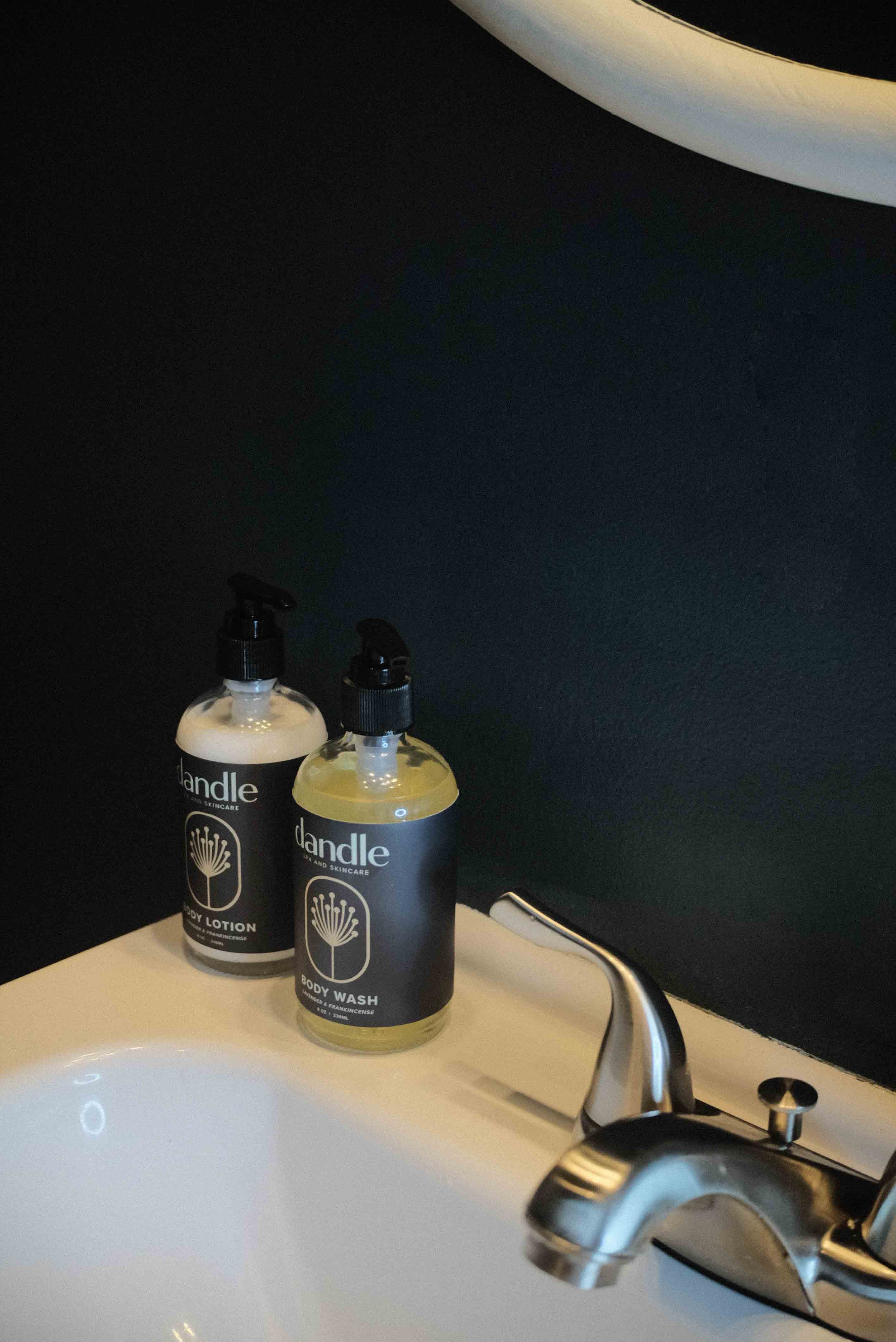 Two bottles of 'Dandle' brand products on a bathroom sink with a dark background. Organic hand soap. 