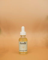 A small glass dropper bottle of Dandle Hair Growth Serum with a white dropper cap, set against a warm beige gradient background.