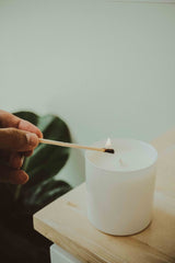 Person lighting a white candle with a match on a wooden surface. Candle is hand-poured in a local boutique in Chattanooga, TN using soy wax and lavender frankincense essential oils.