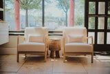 Two beige armchairs with cushions in a room with large windows and a view of trees.
